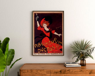 Reklamowy plakat vintage Absinthe Ducros, oprawiony nad komodą z drewna, między roślinami.