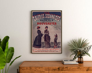 Plakat reklamowy vintage Galeries sedanaises, Nouveautés, oprawiony nad drewnianą komodą, między roślinami.