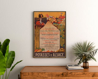 Plakat reklamowy vintage Potasses d’Alsace, oprawiony nad drewnianą komodą między roślinami.