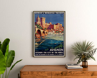 Plakat turystyczny vintage – Avignon, Roman & Mediaeval France, oprawiony nad drewnianą komodą, między roślinami.