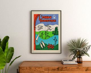 Turystyczny plakat vintage – L’Ain touristique, oprawiony nad drewnianą komodą, między roślinami.