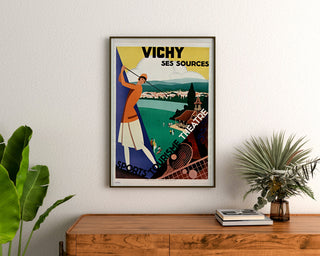 Plakat turystyczny w stylu vintage – Vichy, źródła i turystyka, oprawiony nad drewnianą komodą, między roślinami.