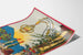 Wintage plakat winiarski – Au bon vin d’Alsace – Hansi, pokazany na zakrzywionej kartce w perspektywie.