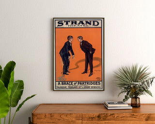 Plakat vintage A Brace of Partridges, Strand Theatre, oprawiony nad drewnianą komodą, między roślinami.