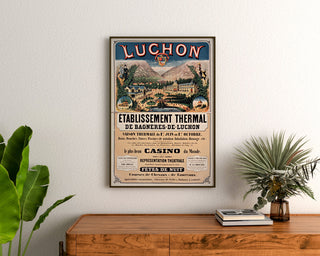 Wintage plakat Bagnères-de-Luchon, uzdrowisko, oprawiony nad drewnianą komodą, między roślinami.