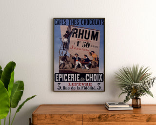 Plakat vintage Épicerie Lefèvre, cafés et chocolats, oprawiony nad drewnianą komodą, między roślinami.