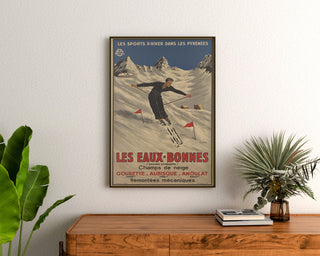 Plakat vintage Les Eaux-Bonnes, sporty zimowe, oprawiony nad drewnianą komodą, między roślinami.