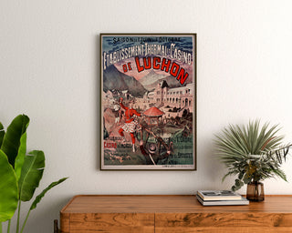Wintage plakat Luchon, kasyno i uzdrowisko, oprawiony nad drewnianą komodą, między roślinami.