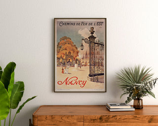 Vintage plakat Nancy, chemins de fer de l’Est, oprawiony nad drewnianą komodą, między roślinami.