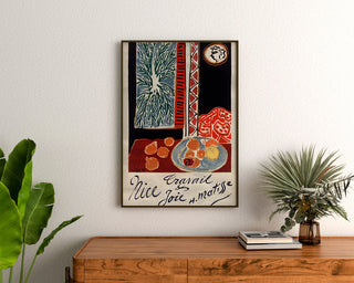 Plakat vintage Nice, Travail et Joie, oprawiony nad drewnianą komodą, między roślinami.
