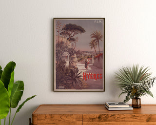 Vintage plakat P. L. M. Hyères, oprawiony nad drewnianą komodą między roślinami.