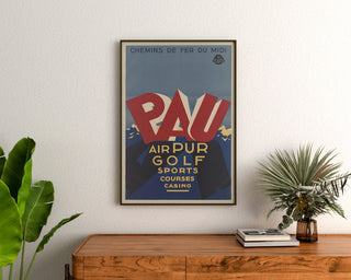 Plakat vintage Pau, golf i sporty, w ramie nad drewnianą komodą, między roślinami.