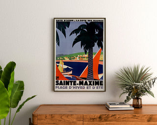 Vintage plakat Sainte‑Maxime, Côte des Maures, oprawiony nad drewnianą komodą między roślinami.